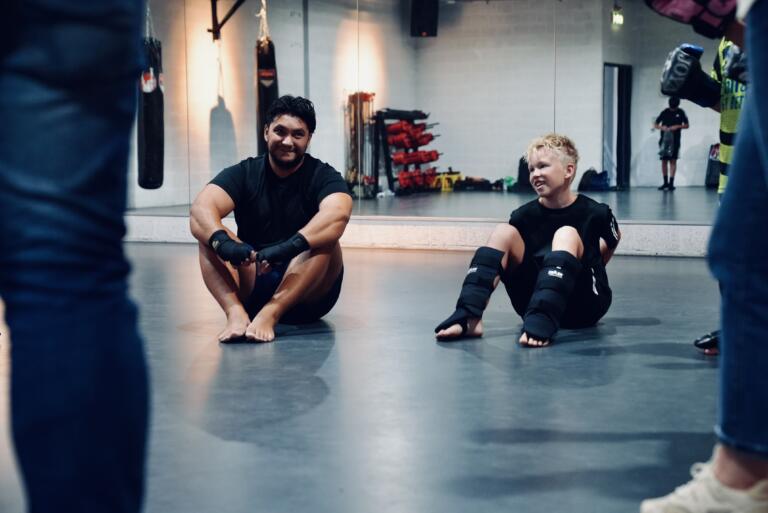 Trainer Ingo van Thaiboxing Center Venlo zit vermoeid maar voldaan op de mat met een lachend kind na de kickboksles
